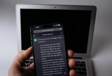 Elde tutulan telefonda açık bir yapay zeka sohbet ekranı, arkada dizüstü bilgisayar