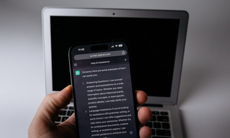 Elde tutulan telefonda açık bir yapay zeka sohbet ekranı, arkada dizüstü bilgisayar